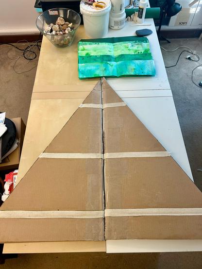 The tree is flattened on the work surface (which is 4 bookcases shoved together) and you can see that there are three lines of cotton tape adhered to the cardboard to join two pieces together. Above it is a pile of green paper, containers of acrylic medium, a bottle of bookbinding glue, tools, and a glass bowl half full of agates.