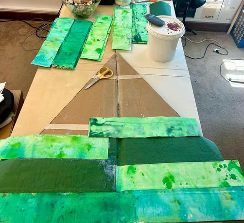 The flat tree is acquiring stripes of green paper! A pair of scissors is sitting on the tree. The pager is spread out above, sorted by color. The lid is on the bucket of gel medium, an acrylic tool rests on top.