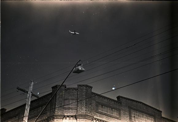 A very old looking photo, despite being brand new, from a 1939 camera. A plane flies over some terrace shop fronts.