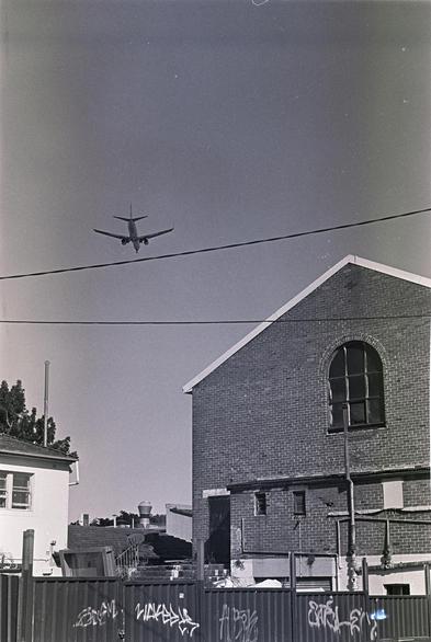 B&W photo of a plane flying over some graffiti-laiden buildings and walls in Sydney's inner west.