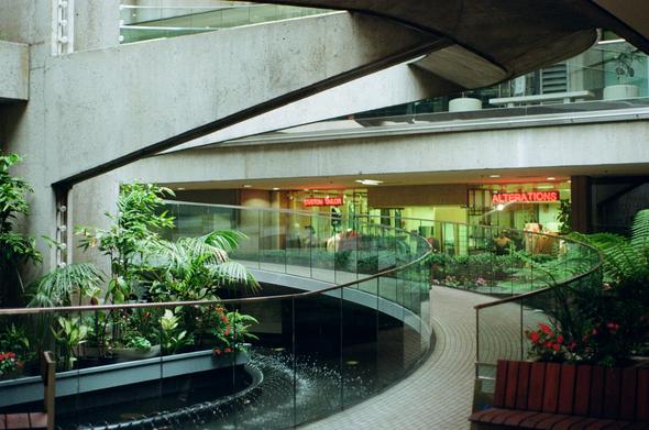 A fountain burbles under a spiral concrete staircase with glass banisters as the neon signs of a tailor beckon in the background.
