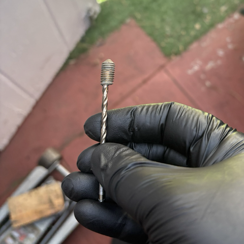 My hand, in a black nitrile glove, holds a drill bit embedded into the end of a bolt with its head broken off.