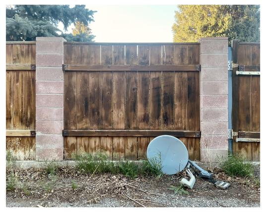 This image shows a wooden fence with alternating vertical planks and a couple of column-like structures made of bricks, enhancing its support and aesthetic. In front of the fence, there is discarded equipment, specifically a disassembled satellite dish and some associated components, which appears to be left abandoned on a patch of dirt and sparse grass. The setting gives a slightly neglected or disused impression.