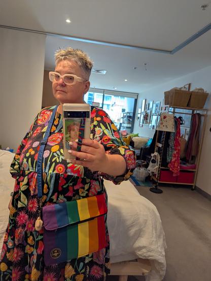Mirror selfie of a person with short hair and white-framed glasses, wearing a brightly coloured dress and a rainbow-striped crossbody bag.