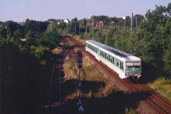 Ein zweiteiliger Dieseltriebwagen der Deutschen Bahn, Baureihe 628 in minttürkiser Lackierung, fährt auf einem eingleisigen Gleis durch eine grüne, leicht hügelige Landschaft. Der Triebwagen ist von schräg oben fotografiert und fährt auf eine Weiche zu, die ihn auf ein Nebengleis führen könnte. Links im Bild steht ein rot zeigendes Hauptsignal. Im Hintergrund sind Bäume, Oberleitungsmasten ohne Fahrdraht und einige Wohnhäuser auf einer Anhöhe zu sehen. Die Szene wirkt sonnig, das Licht erzeugt lange Schatten.