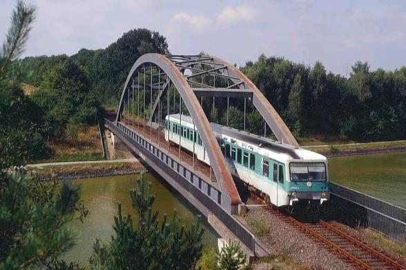 Ein zweiteiliger Dieseltriebwagen der Baureihe 628 in minttürkiser Lackierung fährt auf einer eingleisigen Eisenbahnbrücke über einen Kanal. Die Brücke ist eine moderne Stahlbogenkonstruktion mit auffällig rostfarbenen Bögen, die sich elegant über das Gleis spannen. Der Zug kommt aus einem waldreichen Gebiet und fährt auf den Betrachter zu. Links und rechts unter der Brücke fließt ruhiges Wasser, am Ufer verläuft ein Fußweg. Die Szene ist hell und sonnig, der Himmel leicht bewölkt. Vorne rechts im Bild ragen Tannenzweige ins Bild.