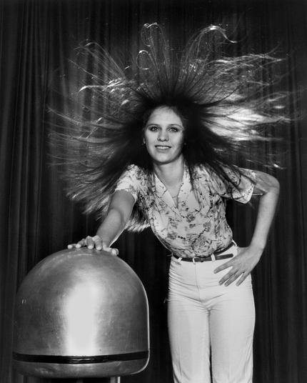 A black and white photo of a young woman with long dark hair, standing on an insulated platform, with one hand on the charge reservoir of a Van de Graaf electrostatic generator.  Her hair is therefore standing almost straight out from her head due to the transfer of charge to each strand of hair, much of which is about half a meter long.

She smiles at the camera, with the other hand on her hip.