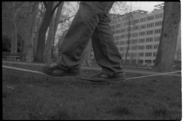 Black and white photography of someone walking on a slack line, in a park in a city. The picture is framed from the knees, centered on the shoes. Slack liner is making a step.