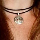 A close-up of a necklace featuring a pendant shaped like a tree, hanging from two black cords. The background shows long, wavy hair.