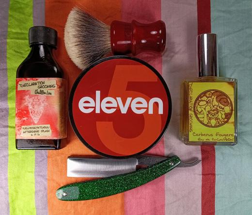 (Le Chat Mistral description)

The image shows a collection of grooming items arranged on a colourful striped cloth. There's a red shaving soap container with the word "eleven" on it, a green straight razor with a shiny blade, a red shaving brush with a white bristle head, and two small bottles. One bottle is labelled "Yuzu/Rose/Patchouli Aftershave Splash" and the other is a yellow perfume bottle labelled "Cerberus Fougere Eau de Parfum". The items are neatly placed, suggesting a grooming or shaving set.