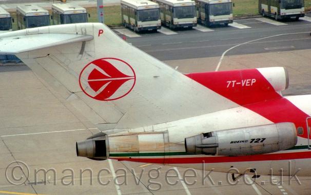 Close-up of the white tail of a 3-engined jet airliner with the engines mounted on the rear fuselage, taxiing from left to right.
There is a thin red and green stripe running along the body, with a wider, red diagonal stripe coming forward from the top of the centre-mounted engine intake, with the white registration "7T-VEP".
The tail has the red outline of a circle with a stylised red flying bird in the middle, and a red letter  "P" right at the top.
The side-mounted engine pods are grey, with black "Boeing 727" text on the sides.
Grey concrete apron fills the background, with a black roadway tacked on to the far side, with blue buses parked side by side along the far edge.