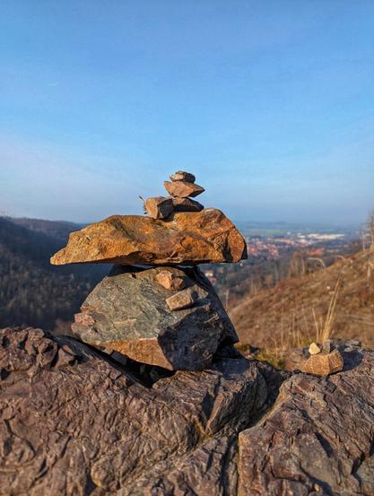 A small cairn of flat, earthy-toned rocks balances precariously on a larger, darker rock. More rocks are scattered at the base and to the right. In the blurred background, a landscape of trees and a distant town are visible under a clear, pale blue sky.