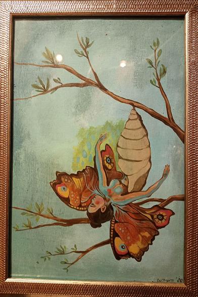 A framed painting, acrylic on canvas. In front of a bluish-gray indefinite background are some twigs with just a few young green leaves. From the top twig, the cocoon of a butterly is suspended. It has just been opened, and a butterfly is hatching. It has the form of a beautiful young woman, red cheeks, brown hair tied to a top knot, arms slightly spread out, breasts exposed, legs still inside the cocoon. She has butterfly wings with a red, yellow and brown pattern. In the bottom right is the artist's signature and a year: "Wayra, '20".