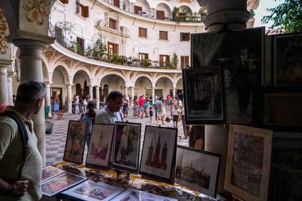 Art prints and postcards on display at a stall in Plaza del Cabildo, Seville, with arched architecture and market visitors in the background.