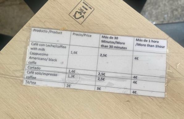 Eine Preisliste für Kaffee- und Teegetränke in spanischer und englischer Sprache mit Angaben zu Produkten wie Café con Leche, Cappuccino, Americano, Cortado, Espresso und Tee sowie deren Preisen und zusätzlichen Gebühren für längere Aufenthaltszeiten.