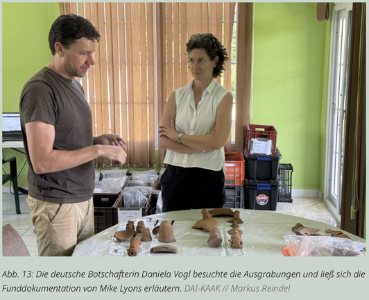 Die deutsche Botschafterin Daniela Vogl informiert sich bei einem Besuch der Ausgrabungen über archäologische Funde. Mike Lyons erläutert die Funddokumentation anhand ausgelegter Keramikfragmente.

Ein Mann (Mike Lyons) steht links an einem Tisch und spricht gestikulierend mit einer Frau (Daniela Vogl), die rechts im Bild mit verschränkten Armen zuhört. Auf dem Tisch liegen zahlreiche archäologische Fundstücke aus Ton, darunter Fragmente von Gefäßen und kleine Figuren. Im Hintergrund stehen beschriftete Kisten und Plastikboxen mit weiteren Fundmaterialien in einem Raum mit grünen Wänden und bodenlangen Jalousien.