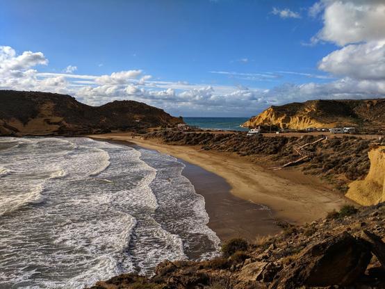 A scenic coastal view showcases a sandy beach meeting the ocean under a partly cloudy sky. The beach is bordered by rocky cliffs on both sides, with gentle waves breaking on the shore. In the distance, a parking space runs parallel to the beach, with cars and campervans parked there. The overall scene captures a tranquil and picturesque seaside setting.
