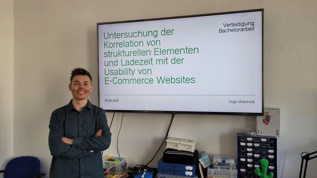Vor einem großen Bildschirm steht links Hugo und lächelt mit verschränkten Armen. Auf dem Bildschirm steht der Titel seine Bachelorarbeit "Untersuchung der Korrelation von strukturellen Elementen und Ladezeit mit der Usability von E-Commerce Webseites".