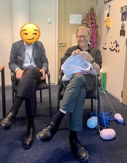 I am sat on a chair in an office, I am knitting something quite big in the round, three balls of coloured yarn are on the floor next to me, a man is on a seat to my right, his face is masked by a smiling side-eye emoji sticker