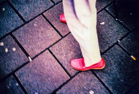 A pair of red shoes worn by some men in the street, wearing also light-coloured pants. The man cannot be seen, the floor is made of concrete tiles where trash is finding shelter. Taken on 35mm Agfa CT Precisa xproed slide film using a Lomography Lc-a camera.