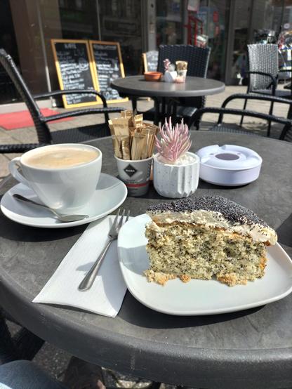 Kaffeetisch mit Deko, Kaffeetasse und einem lockeren Kuchen mit schwarzen Mohnpunkten, der mit weißem Icing bedeckt ist, das wiederum mit einer dicken Schicht aufgestreutem Mohn bedeckt ist.