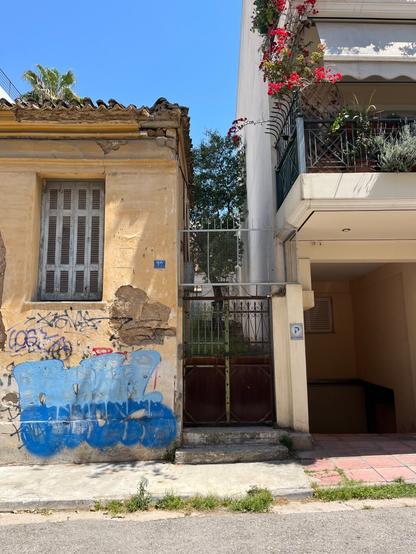 In the Philopappou neighbourhood.
On the left, a one storey neoclassical house in some ruin, with a gate to the side, and on the right part of a three or four storey apartment building.
The older house is a faded sienna yellow with grey wooden shutters and a terracotta tile roof, graffiti under the one window. The wrought iron get has been supplemented with a steel grille above. Just above the roof, the top fronds of a palm tree. 
The newer building is concrete painted white, with balconies the length of the facade, bright with bougainvillea and lavender flowers.
A tree in the depths of the alley between.