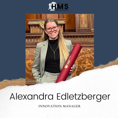 Picture of Alexandra Edletzberger. She has long blond hair, wears glasses, a dark shirt and a beige suit, she smiles.
Under her name it says "Innovation Manager". At the top it says, "HMS - Her Maths Story". Her photo was taken in front of a wooden panel with ornaments and gold.