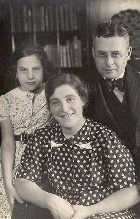 In the center sits a woman wearing a blouse in a pattern of bright stars. Behind her on the left is a young girl, and on the right is a mature man wearing glasses and a suit. Behind them you can see shelves of books.