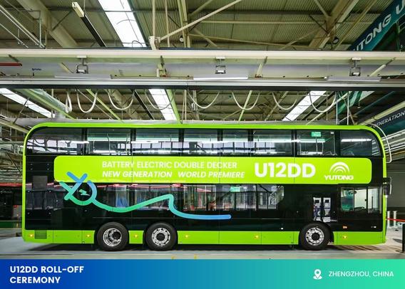 The image features a modern, bright green and black double-decker bus labelled as the U12DD, prominently displayed inside a spacious industrial facility. The upper side of the bus has large windows spanning its length, allowing natural light into the interior. Above the windows, a vibrant green strip runs along the side, bearing bold white text. The bus is positioned centrally with evenly spaced large wheels, emphasising its substantial size. The facility’s ceiling reveals a complex network of exposed beams and lighting, contributing to the industrial atmosphere. In the lower-left corner, a blue strip contains text.