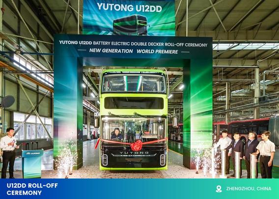 The image shows the launch ceremony of a Yutong U12DD battery electric double-decker bus. The bus, painted in lime green and black, is positioned centrally under a large archway with vibrant green graphics. A folded red ribbon decorates the front of the vehicle. The venue is an industrial hall with high ceilings, exposed metal beams, and large windows on the left-hand side. A group of five formally dressed individuals stand to the right, observing the event. Confetti and sparklers on the ground enhance the celebratory atmosphere.