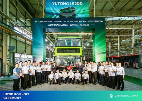 The image depicts a group of people posing in front of a large lime green double-decker bus inside a spacious industrial facility with high ceilings and visible structural beams. The bus stands beneath a vibrant banner and between two green vertical panels. The banner, displaying the model “YUTONG U12DD,” announces the launch of the new battery electric double-decker bus. Formally dressed attendees are gathered in two groups on either side of the vehicle, with some holding a large red ribbon stretched across its front. The setting suggests a ceremonial unveiling. Additional buses in the background indicate a manufacturing or assembly area.