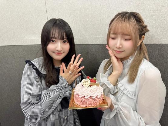Two young women are posing with a beautifully decorated pink and white cake topped with strawberries and a birthday message. One is smiling with her hands held together, while the other has her eyes closed and is gently resting her cheek on her hand, both dressed in soft, feminine outfits.