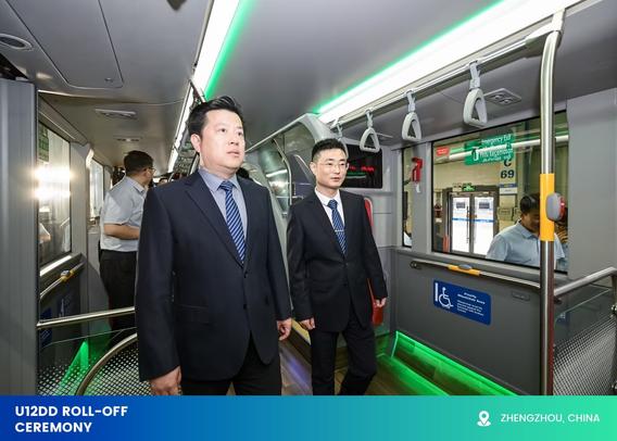 The image shows two men in dark suits and blue striped ties walking inside a modern public transport vehicle, likely a bus or train. The interior features a sleek design with metallic surfaces and a polished wooden floor. Overhead handles hang from the ceiling, and a green light strip lines the top of the compartment. On the right-hand side, a priority seating area is marked by a blue sign with a wheelchair symbol. A window reflects elements of the bright interior. Other passengers are visible in the background, contributing to the clean and professional environment.