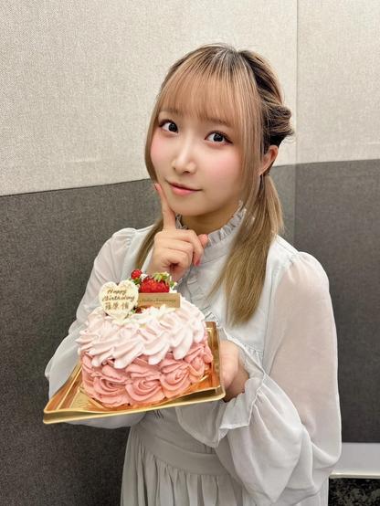 A young woman wearing a white dress is holding a pink rose-patterned cake with a “Happy Birthday” message and strawberries on top. She is posing with a thoughtful expression, touching her chin while looking at the camera.