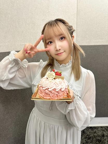 A young woman wearing a white dress is holding a pink rose-patterned cake with a “Happy Birthday” message and strawberries on top. She is posing with a thoughtful expression, doing the peace sign while looking at the camera.