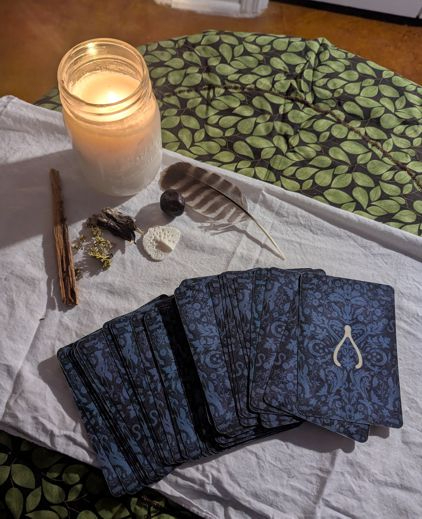 Photo of the Southern Gothic Oracle deck, with a white candle burning. A piece of fat-lighter pine, oak moss, a gator paw, a gator skute, and a feather surround the deck.