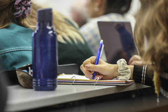 In einem Hörsaal: Studentin schreibt mit einem Stift auf Papier, davor weitere Studierende, ein aufgeklappter Bildschirm, eine Trinkflasche, Mäppchen mit Stiften (Foto: Fabry/Pädagogische Hochschule Karlsruhe)