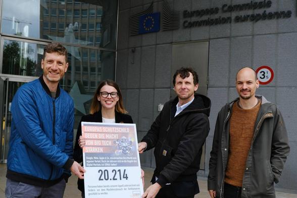 Vier Männer und eine Frau stehen vor der EU-Kommission in Brüssel und halten dabei ein Plakat mit symbolischen 20.214 Unterschriften.