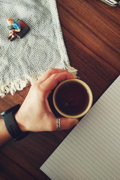 A vertical colour photograph of a small tea cup held in my hand. Around is a blue and white woven cloth with a Tintin figurine drinking tea, and a lined notepad.