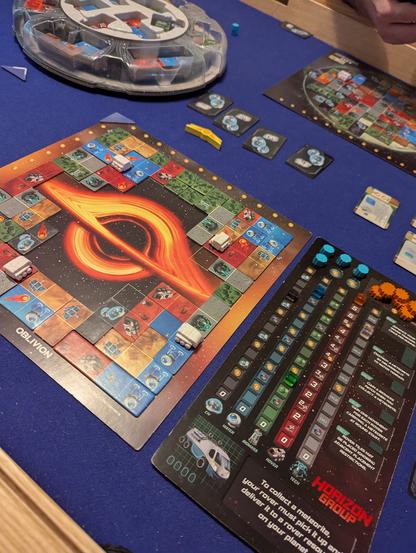A player board for Planet Unknown on the table. It features a black hole with an orange accretion disk. There are polyomino tiles on the board, surrounding the black hole. To the left is a corporation board with lots of tracks.