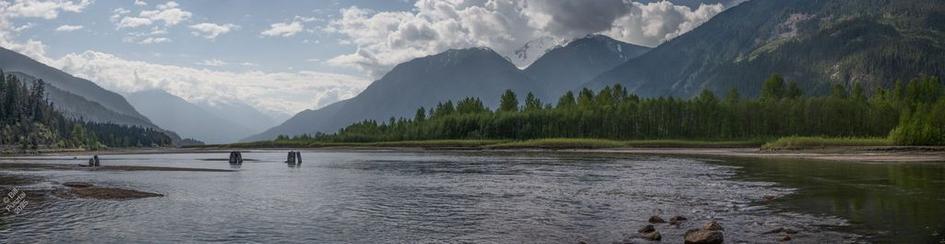 Lillooet Lake. A long thin landscape photo. In the middle is the lake, a widening in the river, shallow and rippling with wind and water flow. Sticking up are several pilings, long since rotted down to stumps. In the distance, successively bluer lighter mountains. Nnearby, wooded slopes, and at river level, short annual vegetation, and behind it stands of alders, all strongly green. The sky is scattered cloudy and sunlight breaks through in rays in places.