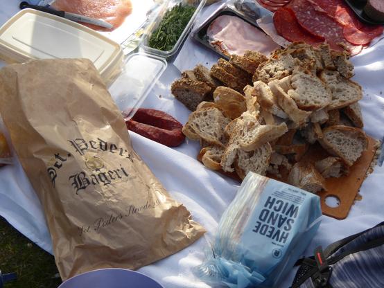 piles of fresh sliced bread, meat, cheese, laid out on a picnic blanket beside bags which read "St. Peder's Bageri" and "HVEDE SANDWICH" (white sandwich bread), waiting for a horde of hungry diners.