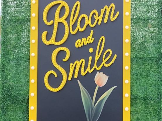 A black sign with yellow script letters and an illustration of a tulip, surrounded by a yellow border. Text reads " bloom and smile."