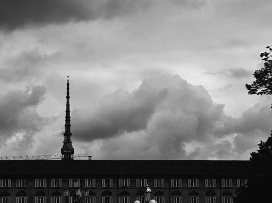 The photo is in black and white and shows a building called mole antonelliana, set against a cloudy sky.  and there's a tree on the right side. The sky is filled with large clouds.