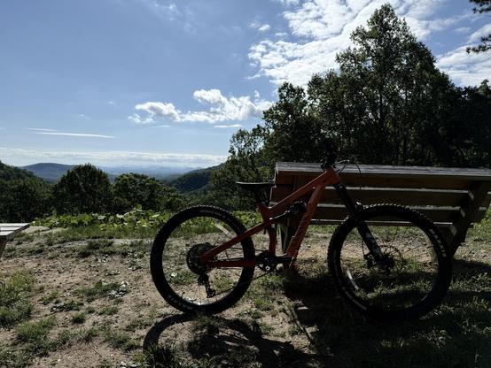 A mountain bike is positioned in the foreground, with a scenic view of rolling hills and a blue sky in the background. A wooden bench is nearby, surrounded by greenery.