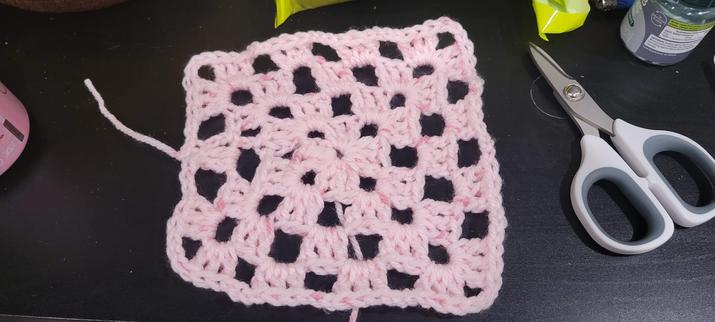 A granny square laying flat on a black table. The "square" is lopsided, and you can see many parts where the tension in the stitches was too loose. The piece is made with a very light pink, almost white, acrylic yarn.