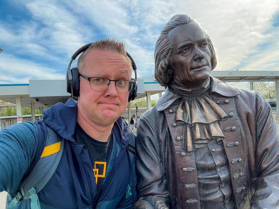 A person wearing headphones poses for a selfie next to a bronze statue of a historical figure, set against a clear blue sky with some clouds.