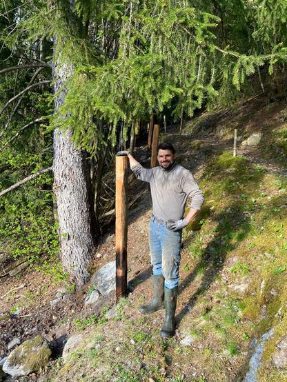A young man is standing and smiling near a wooden fence post, with one hand resting on top of a larch fence post. A large spruce tree is visible on the left side of the photo.