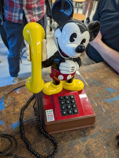 Tastentelefon in rot mit schwarzen Tasten auf einer Werkbank, darauf eine Mickey Mouse Figur, die den gelben Hörer festhält