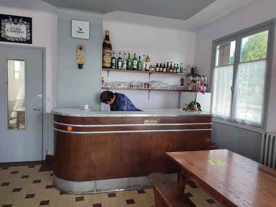 À gauche une porte qui mène vers le logement  de Arlette. Arlette derrière le bar en bois. À droite une fenêtre avec des vieux rideaux. Au premier plan une table en bois